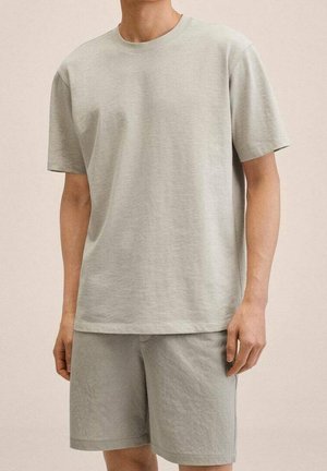 Mężczyzna w prostym jasnoszarym t-shircie z krótkim rękawem i pasujących szortach, stojący na neutralnym tle.