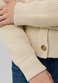 Cardigan tricoté de couleur beige clair avec des poignets côtelés. Il présente une fermeture à boutons à l'avant avec un bouton rond et transparent. Détails de tissu texturé visibles.