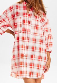 Robe chemise oversized à carreaux rouges et blancs, avec un col en V, un boutonnage sur le devant et deux poches poitrine. Tissu léger à carreaux.