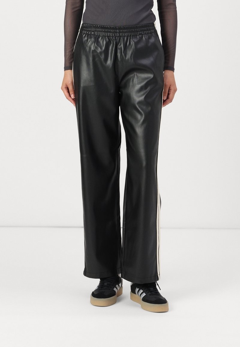 Personne portant un pantalon noir en simili cuir avec une taille élastique et des bandes blanches sur les côtés, associé à des baskets noires et blanches.