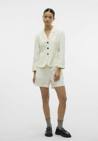 Blazer avorio con revers a punta, bottoni neri, due tasche frontali; abbinato a pantaloni corti coordinati. Il modello indossa calze grigie e mocassini neri.