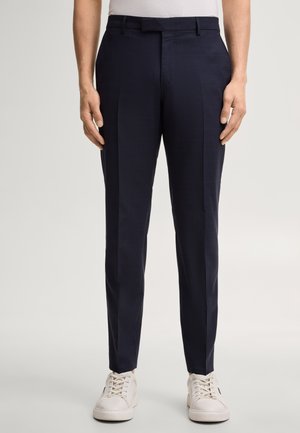 Man draagt slim-fit marineblauwe nette broek en witte sneakers tegen een effen achtergrond, handen ontspannen langs de zijkanten.