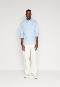Camisa azul claro de botones con mangas largas, combinada con pantalones anchos blancos y zapatillas blancas, sobre un fondo sencillo.