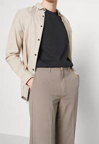 Béžová lněná košile s knoflíky, nošená přes černé tričko. Kombinováno s béžovými kalhotami na míru se bočními kapsami a hladkou texturou.