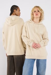 Beige hooded sweatshirt in a relaxed fit, featuring a drawstring, long sleeves, and side pockets. Worn with loose denim pants.