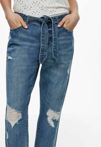 Vaqueros de denim azul claro con cintura ajustada, que presentan parches desgastados y un diseño de pierna recta. Textura suave con un aspecto ligeramente usado.