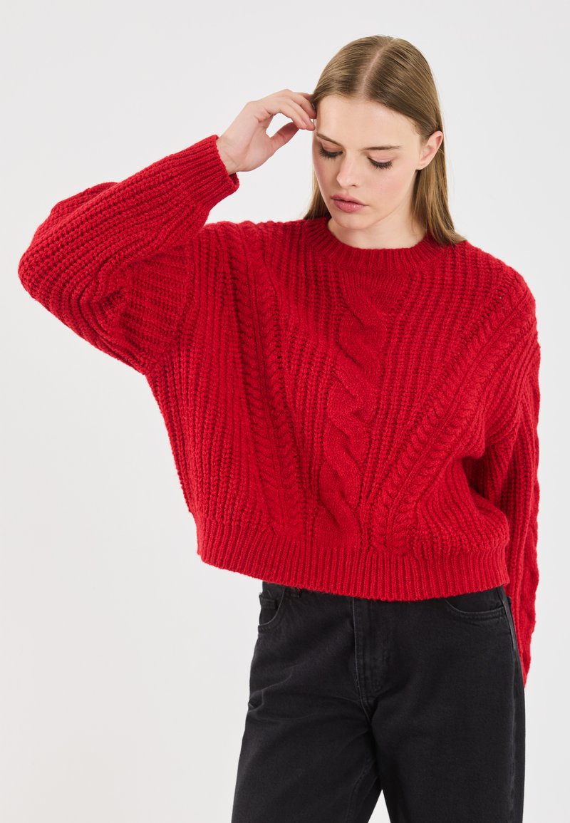 Junge Frau, die ihr Haar mit der Hand berührt, trägt einen übergroßen roten Zopfpullover und schwarze Hosen vor einem schlichten Hintergrund.