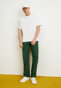 Tricou alb din bumbac, pantaloni verzi, teniși albi și o șapcă albă. Acest ansamblu are o croială lejeră și un design minimalistic.