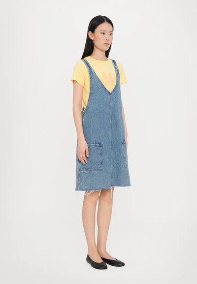 Robe salopette en denim avec un décolleté en V, des poches latérales et un ourlet effiloché. Portée par-dessus une chemise jaune avec un graphique de soleil. Chaussures noires plates.