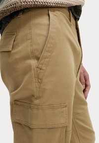 Beige cargo kalhoty s hladkým materiálem, bočními kapsami a detailním šitím podél švu. Kalhoty jsou navrženy s rovnými nohavicemi.