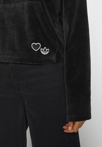 Svart, sammetslen hoodie med en framficka, prydd med en vit broderad hjärta och tre ränder logga på vänster sida. Svarta jeans synliga.