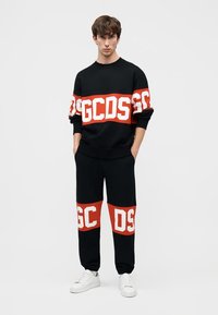 Jaunas vyras stovintis su rankomis kišenėse, dėvintis juodą džemperį ir kelnes su ryškiomis raudonomis ir baltomis GCDS logotipo juostomis, bei baltais sportbačiais.