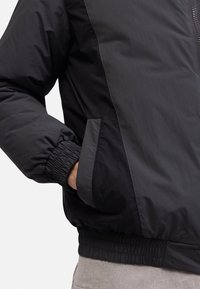 Black jacket with a smooth, matte finish. Features elastic cuffs, a zippered front, and side pockets with contrasting trims.
