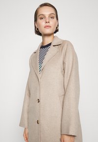 Beige wollen jas met een gestructureerd ontwerp, geknipte revers, sluiting met twee knopen en zijzakken. Beschikt over een zachte textuur en een oversized vorm.