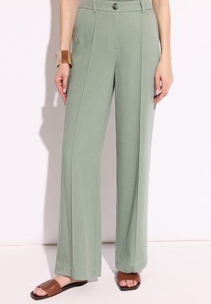 Jasnozielone spodnie o szerokich nogawkach z przednią kantą, noszone z brązowymi sandałami typu slide i masywną bransoletką na lewym nadgarstku.