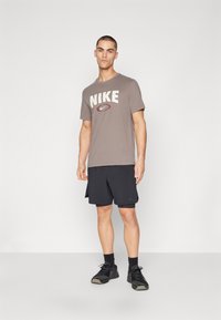 Grå Nike t-skjorte med fet logo, kombinert med svarte treningsshorts. Modellen har på seg svarte joggesko og ankelstrømper, stående foran en ensfarget bakgrunn.