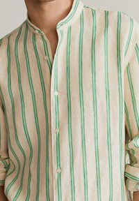 Leinenhemd mit beigegrund und vertikalen grünen Streifen, mit Stehkragen und durchgehender Knopfleiste, lange Ärmel hochgekrempelt.