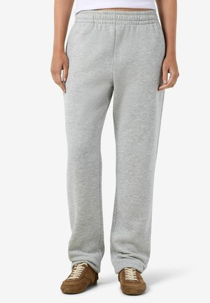 Personne portant un pantalon de survêtement gris clair avec taille élastique et des chaussures décontractées marron à lacets, debout devant un fond uni.