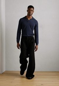 Suéter navy con cuello en V y solapa, combinado con pantalones anchos negros. El modelo se encuentra de pie sobre un suelo de madera frente a una pared de color gris claro.