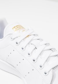 Baltas ādas sneakers ar perforētu apdari, plakaniem baltajiem šņorēm un zelta logo tagu uz valkāšanas vietas. Gluda tekstūra un zema profila dizains.