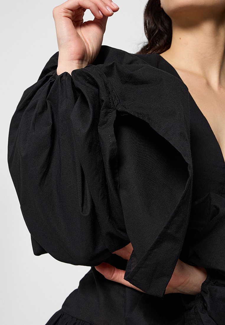 Schwarzes Kleidungsstück mit Puffärmeln aus strukturiertem Stoff, das geraffte Ärmel und einen tiefen Ausschnitt aufweist. Sichtbare Hand und Handgelenk in einer dezenten Pose.