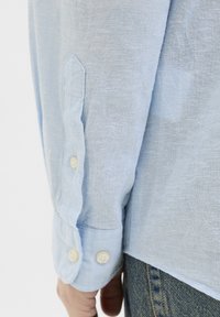 Camisa de lino azul claro con mangas largas, que presenta un puño abotonado y una tela ligeramente texturizada. Los botones visibles son blancos y redondos.
