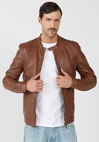 Braune Lederjacke mit einem Reißverschluss vorne, rundem Kragen und gerippten Akzenten auf den Schultern. Getragen über einem weißen Hemd mit Jeans.