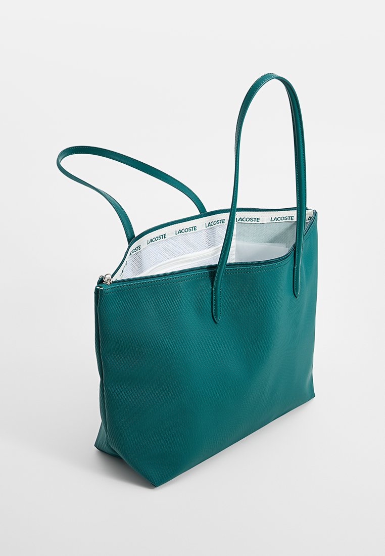 Bolso tote sintético en color azul verdoso con dos asas largas, exterior texturizado y un interior blanco que presenta un estampado de logotipos repetidos a lo largo del borde superior.