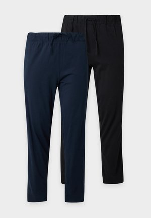 Hombre de pie descalzo con una camiseta gris de manga corta y pantalones deportivos azul oscuro frente a un fondo claro y liso.