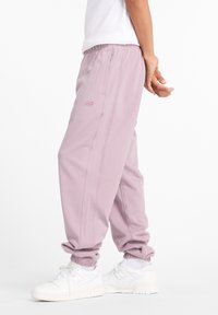 Hellviolette Jogginghosen mit elastischem Bund und Seitentaschen. Sie haben eine schlanke Passform, zulaufende Beine und ein dezentes Logo an der Seite. Weiße Turnschuhe.