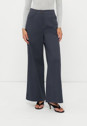 Mujer que lleva pantalones oscuros de pierna ancha, camiseta gris de manga larga y sandalias negras de tacón, de pie frente a un fondo blanco liso.