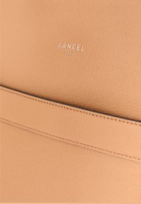 Lancel Paris handtas van zacht beige gestructureerd leer, met subtiele stiksels en een aangebracht logo-detail, met een soepel, strak ontwerp.