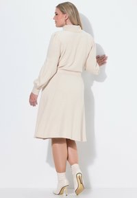 Abito beige a maniche lunghe con colletto e vita affusolata, che si apre in una gonna lunga fino al ginocchio. Indossato con stivaletti bianchi alla caviglia con dettagli dorati.