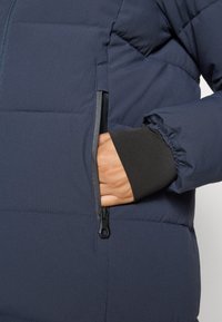 Marineblå dunjakke med horisontalt quiltet mønster, glidelåslomme, elastiske mansjetter og kontrastfarget svart innerfôr. Smoother tekstur.