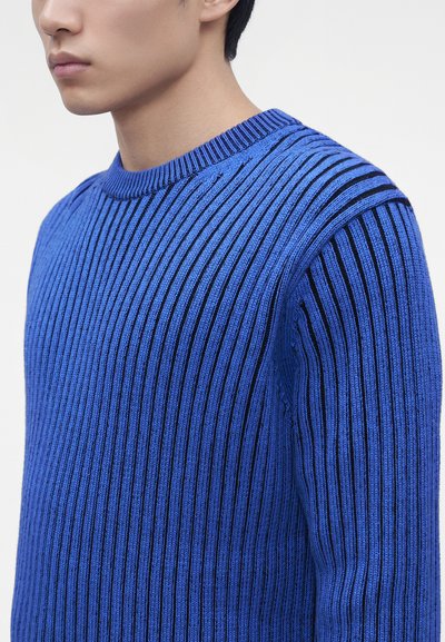 Gros plan d'une personne portant un pull tricoté côtelé bleu avec des rayures verticales noires, montrant le haut du torse et le cou.