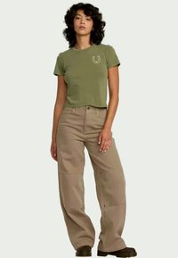 Femme aux cheveux bouclés portant un t-shirt cropped vert olive avec un motif fer à cheval et un pantalon beige à jambes larges, avec des bottes noires.