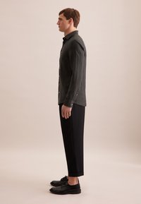 Camicia nera a maniche lunghe con colletto a bottoni, abbinata a pantaloni neri aderenti e scarpe nere, modello mostrato di profilo su sfondo chiaro.