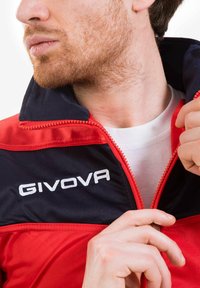 Giacca rossa e blu presenta una zip frontale, colletto alto e logo ricamato "GIVOVA". Il tessuto appare liscio con un design sportivo.