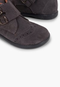 Zapatos de ante gris con puntera redondeada, cierre de doble hebilla, perforaciones decorativas y suela de goma negra.