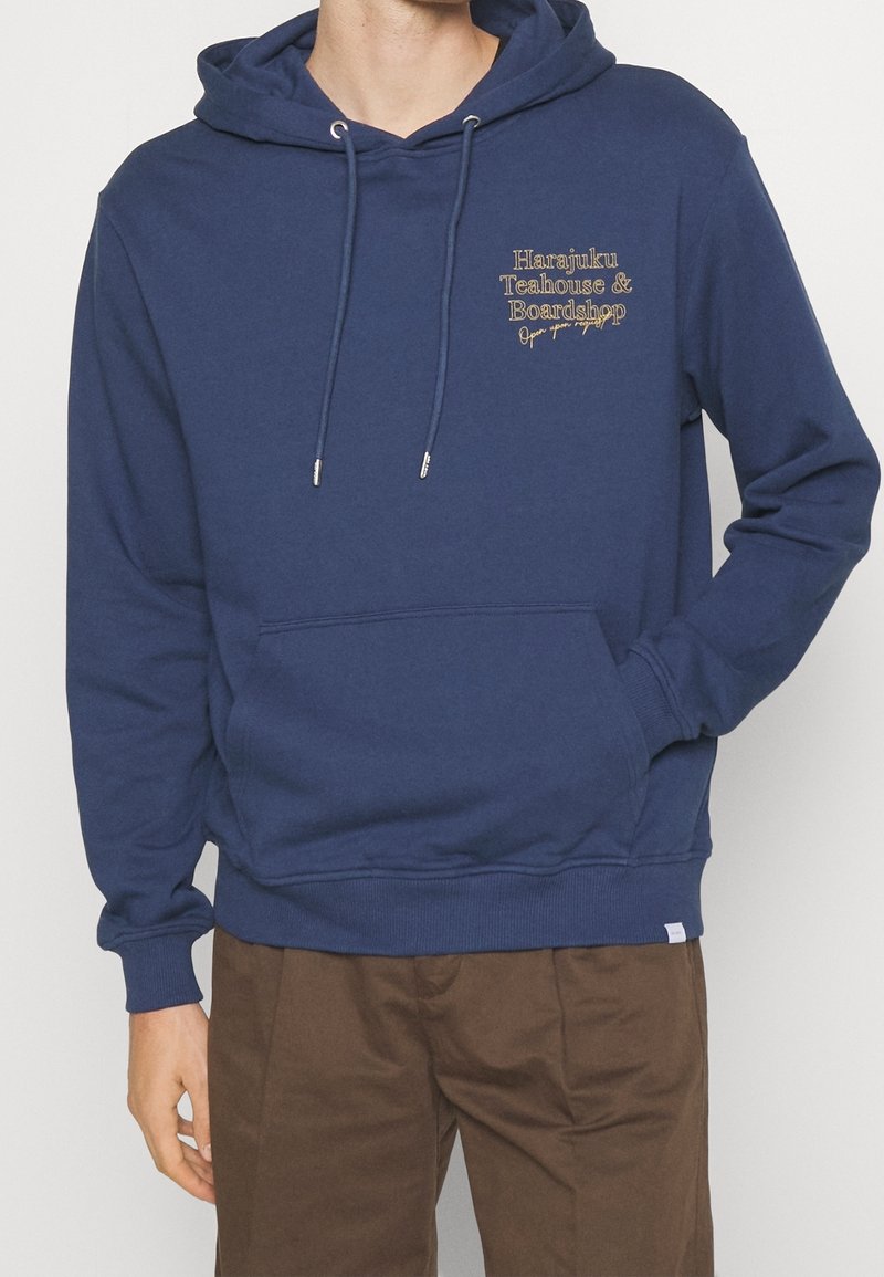 Uomo che indossa un hoodie blu navy con la scritta "Harajuku Teahouse & Boardshop" sul petto e pantaloni marroni, in piedi con una mano in tasca.