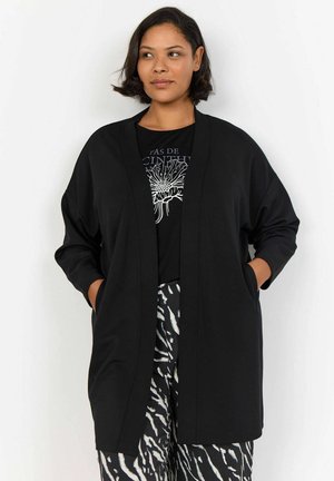 Veste de style kimono noire avec des manches longues, à l'avant ouvert, et deux poches latérales, faite d'un tissu lisse ; portée sur un t-shirt graphique noir.
