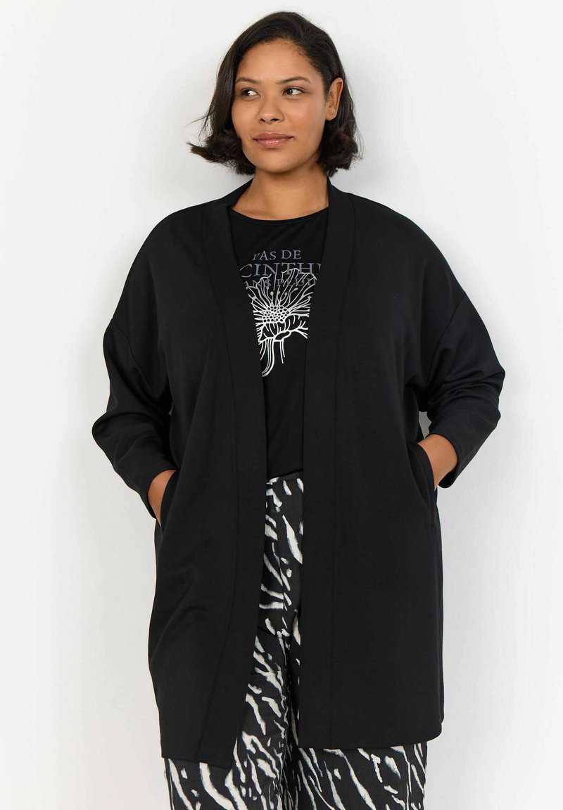Musta kimono-tyylinen takki, jossa on pitkät hihat, avonainen etuosa ja kaksi sivutaskua, valmistettu sileästä kankaasta; päällä graafinen musta t-paita.