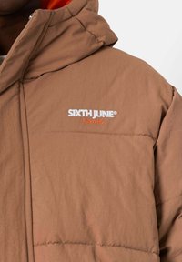 Bruine gewatteerde jas met een capuchon, voorzien van geborduurde tekst "SIXTH JUNE PARIS" in wit en oranje. Gladde textuur met doorgestikte details.