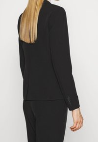 Blazer nero su misura con vestibilità slim, maniche lunghe, spalline sottili e cinque bottoni decorativi sui polsini. Tessuto liscio.