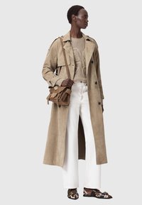 Trenci bej din piele întoarsă peste un pulover bej tricotat, asortat cu pantaloni albi cu croială largă și mocasini cu model, purtând o geantă maro de tip crossbody.