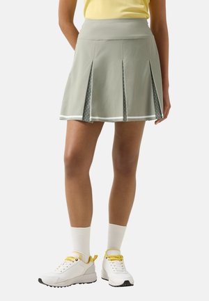 Frau trägt einen hellgrauen Faltenrock mit gemusterten Einsätzen, weiße Socken, weiße Turnschuhe mit gelben Schnürsenkeln und ein gelbes Oberteil.