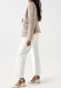 Femme portant une veste en tweed texturée, un pantalon blanc taille haute à jambes larges, et des chaussures Mary Jane beige à talons épais sur un fond blanc.