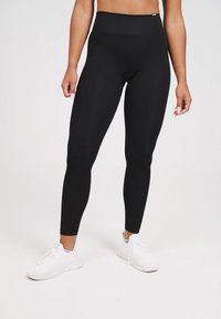Zwarte hooghangende leggings van een gladde, elastische stof. Strak ontwerp en naadloze constructie. Gemaakt in combinatie met witte sport schoenen.