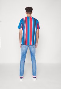 Joven de pie frente a una pared, vistiendo una camiseta de rayas verticales en azul, rojo y blanco, pantalones vaqueros de color azul claro y zapatillas blancas.