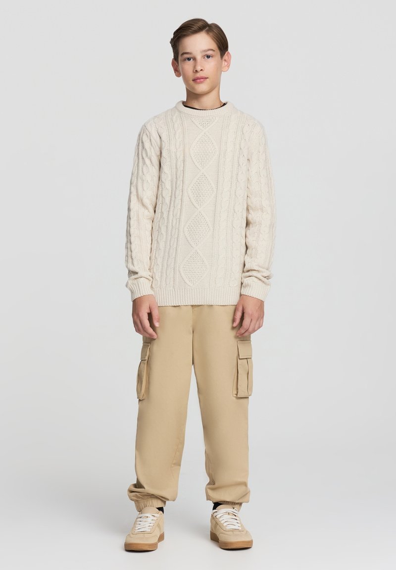 Maglione crema a trecce con motivo a rombi, abbinato a pantaloni cargo color sabbia con tasche laterali e sneakers beige.